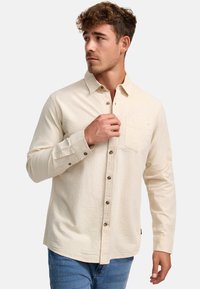 Chemise beige texturée à boutons avec une poche et des boutons marron, associée à un jean bleu. Le tissu présente un motif subtil et une douceur.