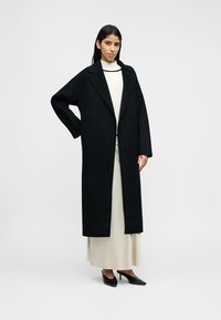 Manteau long en laine noire avec col cranté, silhouette droite et poches latérales, porté sur une robe crème et des escarpins noirs.