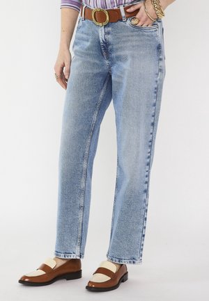 Personne portant un jean droit bleu clair avec une ceinture marron, des mocassins marron et crème, et des bijoux en or à une main.