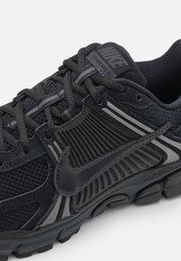 Nike Volero 5 kedas melnā krāsā ar sietiņa augšdaļu, pelēkiem akcentiem, gumijas zolēm un mīkstinātu starpzoli. Iespējas ietver izteiktu logotipu un fakturētu dizainu.