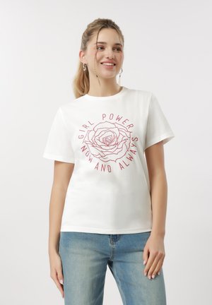 Noor naine naeratab, kandes valget t-särki, millel on punane roos ja tekst "Girl Power Now and Always", ning hele sinine teksapüksid.