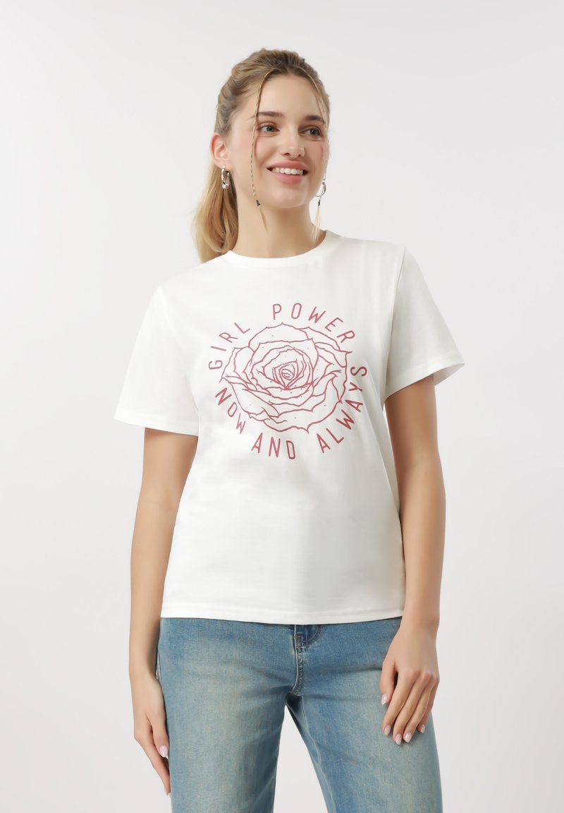 Νεαρή γυναίκα που χαμογελά, φορώντας λευκό μπλουζάκι με κόκκινο τριαντάφυλλο και το κείμενο "Girl Power Now and Always," συνδυασμένο με ανοιχτόχρωμα μπλε τζιν.