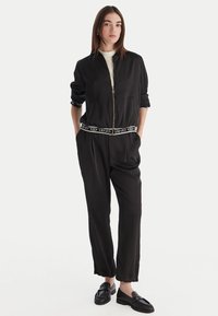Tuta nera con giacca bomber con zip e vita elasticizzata. Tessuto morbido, vestibilità rilassata, con logo dettagliato sulla vita.