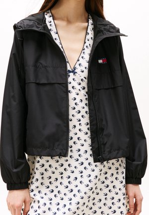 Vrouw die een zwart jasje met rits draagt over een witte jurk met een marineblauw vogelpatroon en een klein Tommy Jeans-logo op de borst van het jasje.