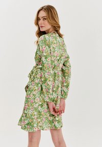 Kvetinové zelené šaty s priliehavým pásom, dlhými rukávmi a rozšíreným spodným okrajom. Vzor obsahuje ružové a biele kvety na listovom pozadí.
