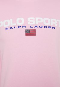 Camisa rosa con "POLO SPORT" en blanco, "RALPH LAUREN" en azul y una pequeña bandera estadounidense debajo del texto en la zona del pecho.