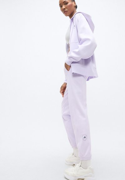 Σετ φόρμας λιλά που περιλαμβάνει ζακέτα με φερμουάρ και παντελόνια που στενεύουν στο κάτω μέρος, κατασκευασμένο από απαλό υλικό. Περιλαμβάνει λογότυπο στα παντελόνια και λευκά αθλητικά παπούτσια.