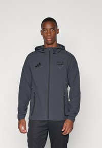 Grå hettejakke laget av lett materiale, med glidelås foran, to glidelåslommer, og svart Adidas-logo og Arsenal-emblem.