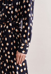 Vestido de marinero con grandes lunares blancos y naranjas, detalle de cinturón en la cintura y puños abotonados, que presenta una textura de tela suave y lisa.