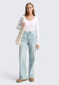 Langärmliges weißes Oberteil kombiniert mit hellblauen weit geschnittenen Jeans und beigen Plateauschuhen, während eine beige Tragetasche getragen wird. Minimalistisches Design, lässiger Stil.