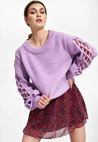 Lila gebreid sweater met brede, gevlochten mouwen, gecombineerd met een korte, bloemenpatroon rok met een zwarte achtergrond en kleurrijke bloesems.