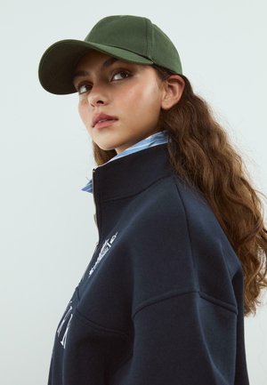 Sweatshirt bleu marine avec un col haut et une fermeture éclair, orné de broderies blanches. Une casquette verte est portée de travers, recouvrant partiellement des cheveux bouclés.