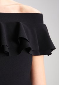 Czarna sukienka odkrywająca ramiona z falbaną przy dekolcie. Gładki materiał, strukturalny kształt i miękki detal przy ramieniu.