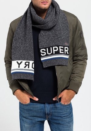 Homme portant une veste bomber vert olive, un pull foncé, un jean bleu et une épaisse écharpe grise avec les textes "SUPER" et "DRY".
