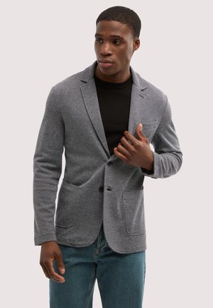 Mann trägt einen strukturierten grauen Blazer über einem schwarzen Hemd und blauen Jeans und richtet den Blazer mit einer Hand.