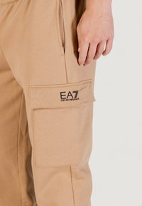 Pantaloni cargo beige realizzati in tessuto morbido, con una tasca laterale con patta e un logo ricamato nella zona della coscia.