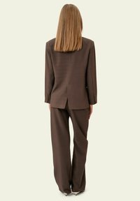 Mujer con cabello rubio lacio, vistiendo un traje holgado marrón con pantalones de pierna ancha y zapatos de tacón alto, de pie de espaldas.