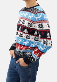 Homme portant un pull festif avec des motifs de rennes, de flocons de neige et de sapins de Noël dans des tons de rouge, bleu, blanc et noir, associé à un jean bleu.
