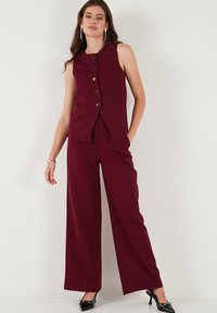 Bordový bezrukávník s velkými knoflíky a bočními kapsami, v kombinaci s širokými kalhotami ze stejné látky a barvy. Černé podpatky dotvářejí outfit.