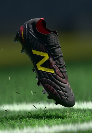 Schwarzer Fußballschuh mit gelbem "N"-Logo und roten Akzenten, der Gras auf einem grünen Spielfeld nahe einer weißen Linie aufwirbelt.