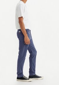 Tmavě modré chino kalhoty s úzkým střihem, bavlněná látka, pět kapes a nášivka vzadu. Kombinovány s bílým tričkem s krátkým rukávem.