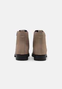 Par de botas de tobillo beige con suelas negras, vistas desde atrás, con el logo de TOMS en las lengüetas traseras sobre un fondo blanco.