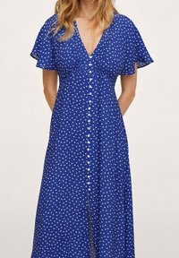 Sininen pilkullinen mekko, jossa on lyhyet lepattelevat hihat, V-pääntie, etupainonappi sulkeminen ja halkio edessä liikkumisen helpottamiseksi.