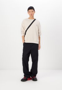 Felpa beige con design ricamato, pantaloni cargo neri con tasche laterali e sneakers nere con dettagli rossi.