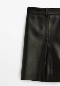 Jupe taille haute en simili cuir noir avec fente centrale à l'avant et passants de ceinture, présentée sur un fond blanc.