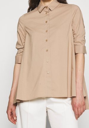 Camicia beige a maniche lunghe con colletto a punta, maniche arrotolate e vestibilità ampia. Presenta bottoni marroni e un tessuto liscio e leggero.
