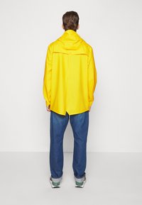Person iført en lys gul regnjakke med hætte, blå oprullede jeans og hvide sneakers, der står vendt mod en ensfarvet lys baggrund.
