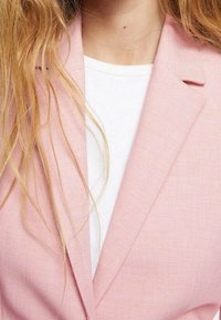 Jasnoróżowa marynarka z teksturowanej tkaniny, z klapami typu notch. Noszona na białej koszuli, z częściowo widocznymi miękkimi, falującymi włosami.