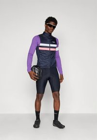 Άνδρας με μωβ μακρυμάνικη μπλούζα, ναυτικό γιλέκο ποδηλασίας, μαύρες βερμούδες ποδηλασίας, κρατώντας μαύρο κράνος, φορώντας γυαλιά ηλίου και παπούτσια ποδηλασίας.