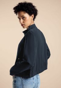 Marineblaues, kurzes Jackett mit Stehkragen, langen Ärmeln und elastischem Saum; glatte Textur; kombiniert mit hellblauer Jeans.