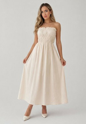 Femme portant une robe bustier écrue longue jusqu'aux chevilles, tenant les côtés de la jupe, posant devant un fond clair uni.