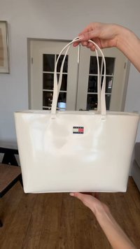 Κρεμ λευκή τσάντα Tote Tommy Jeans κρατημένη από δύο χέρια, με διπλές λαβές και λογότυπο στο λείο ύφασμα.
