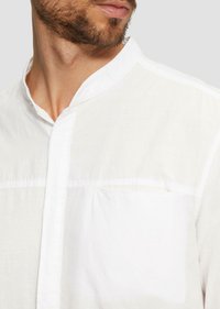 Witte, knooploze, gekraagde blouse van een gestructureerde stof met een borstzak, voorzien van een subtiele horizontale naad aan de voorkant.