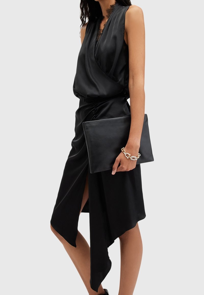 Femme portant une robe portefeuille noire en satin sans manches tenant une grande pochette noire avec un bracelet chaîne dorée à son poignet.