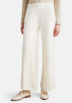 Woman wearing white wide-leg trousers, beige long-sleeve top, beige socks, and cream-colored loafers standing on a neutral background.