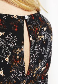 Schwarzes Blumen-Kleid mit einer Knopfverschluss hinten. Es hat kleine weiße und goldene Blumenmuster sowie einen Schlüsselaugen-Ausschnitt am Halsausschnitt.