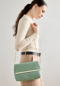 Woman in beige top and pants holding a green handbag with a gold chain strap over her shoulder against a plain background.