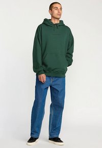 Zelen pulover s kapuco in sprednjim žepom, kombiniran z modrimi kavbojkami s ravnimi hlačnicami in črnimi supergami. Enostaven dizajn, bombažni material.