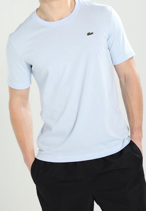 Mand iført en lyseblå T-shirt med korte ærmer med et lille grønt krokodillelogo og sorte bukser, hænderne i lommerne.