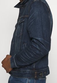 Dark blue denim jacket with a front button closure, two chest pockets, and a fitted design. Features prominent stitching and a collar.