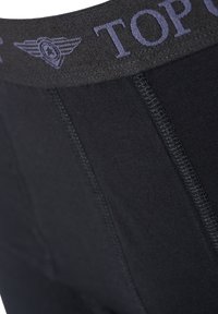 Mustat potkupöksyt, joissa on sileä kangas ja leveä vyötärönauha kohokuvioidulla tekstillä ja logolla. Mallissa on litteät saumat mukavuutta varten.