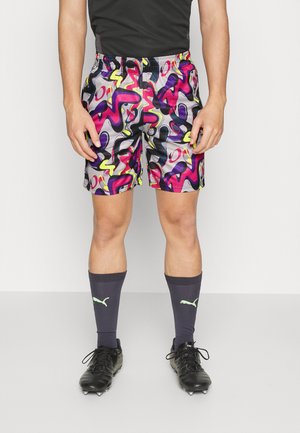 Athlète portant un short coloré à motif abstrait, des chaussures à crampons noires et des chaussettes Puma foncées, debout devant un fond clair uni.