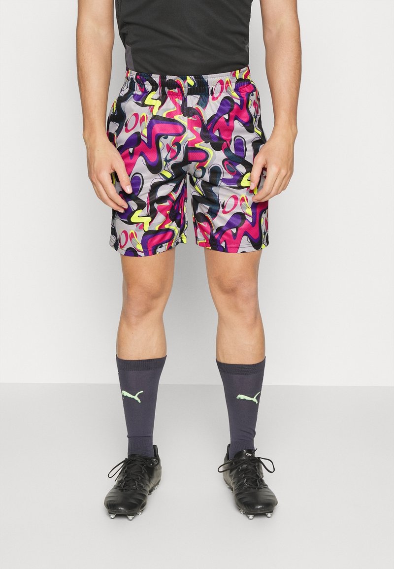 Athlète portant un short coloré à motif abstrait, des chaussures à crampons noires et des chaussettes Puma foncées, debout devant un fond clair uni.