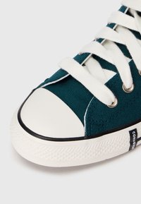 Sneaker in tela teal con lacci bianchi, punte di gomma e suola testurizzata con un accento nero. Etichetta del marchio visibile sul lato.