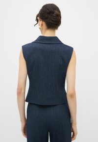 Ärmlös mörk denimväst med spetsig krage, figursydd design och en ren finish vid fållen. Noterbar textur med en subtil lyster.