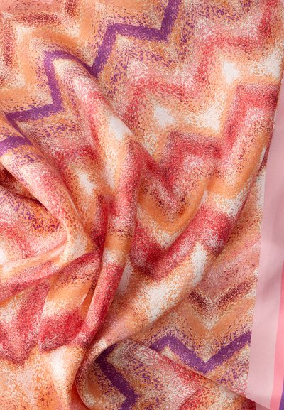 Μαλακό ύφασμα με υφασμένους ζιγκ ζαγκ σχεδιασμούς σε αποχρώσεις του πορτοκαλί, κόκκινου, μοβ και λευκού, κρεμασμένο με ορατές πτυχώσεις σε ροζ φόντο.